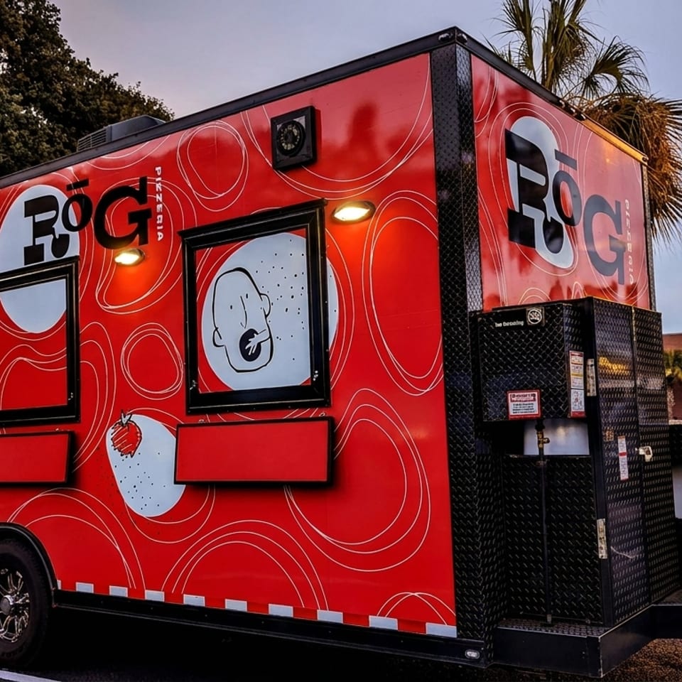 Red food truck with "ROG Pizzeria" logo and abstract white designs parked at dusk.