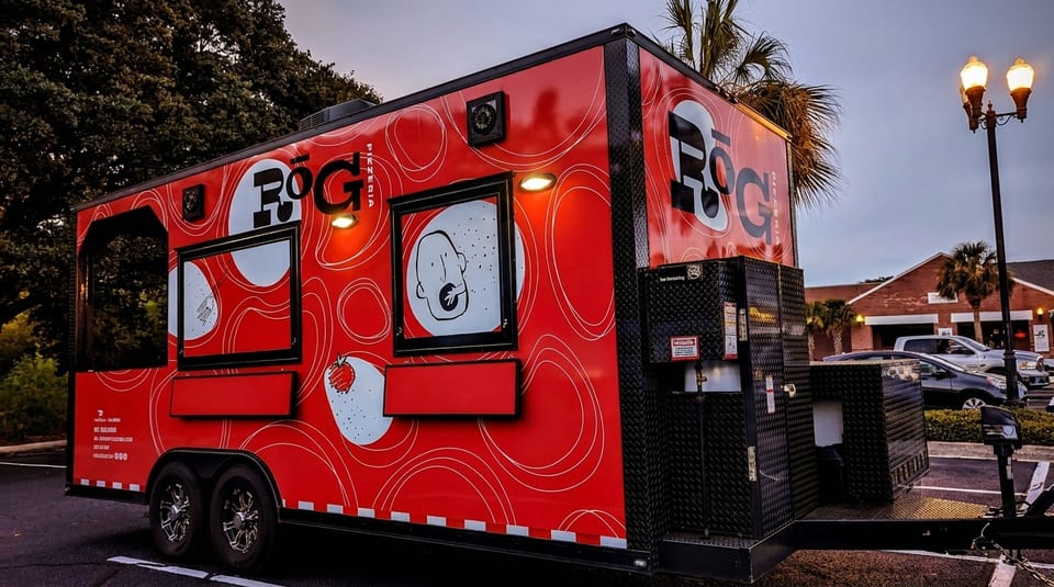 Red food truck with "ROG Pizzeria" logo and abstract white designs parked at dusk.