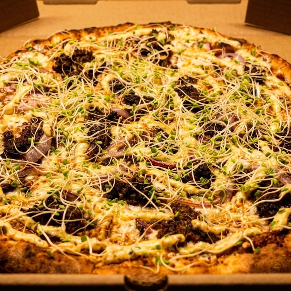 A whole pizza in a cardboard box, topped with cheese, dark meat, red onions, and sprouts.