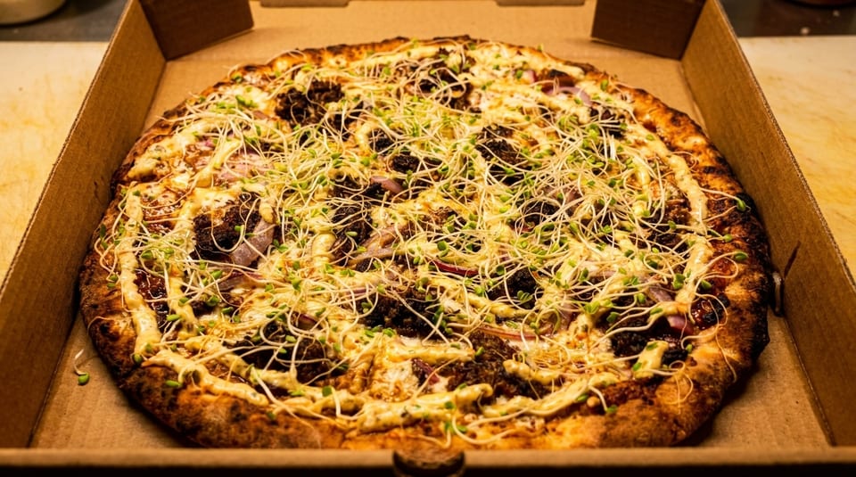 A whole pizza in a cardboard box, topped with cheese, dark meat, red onions, and sprouts.
